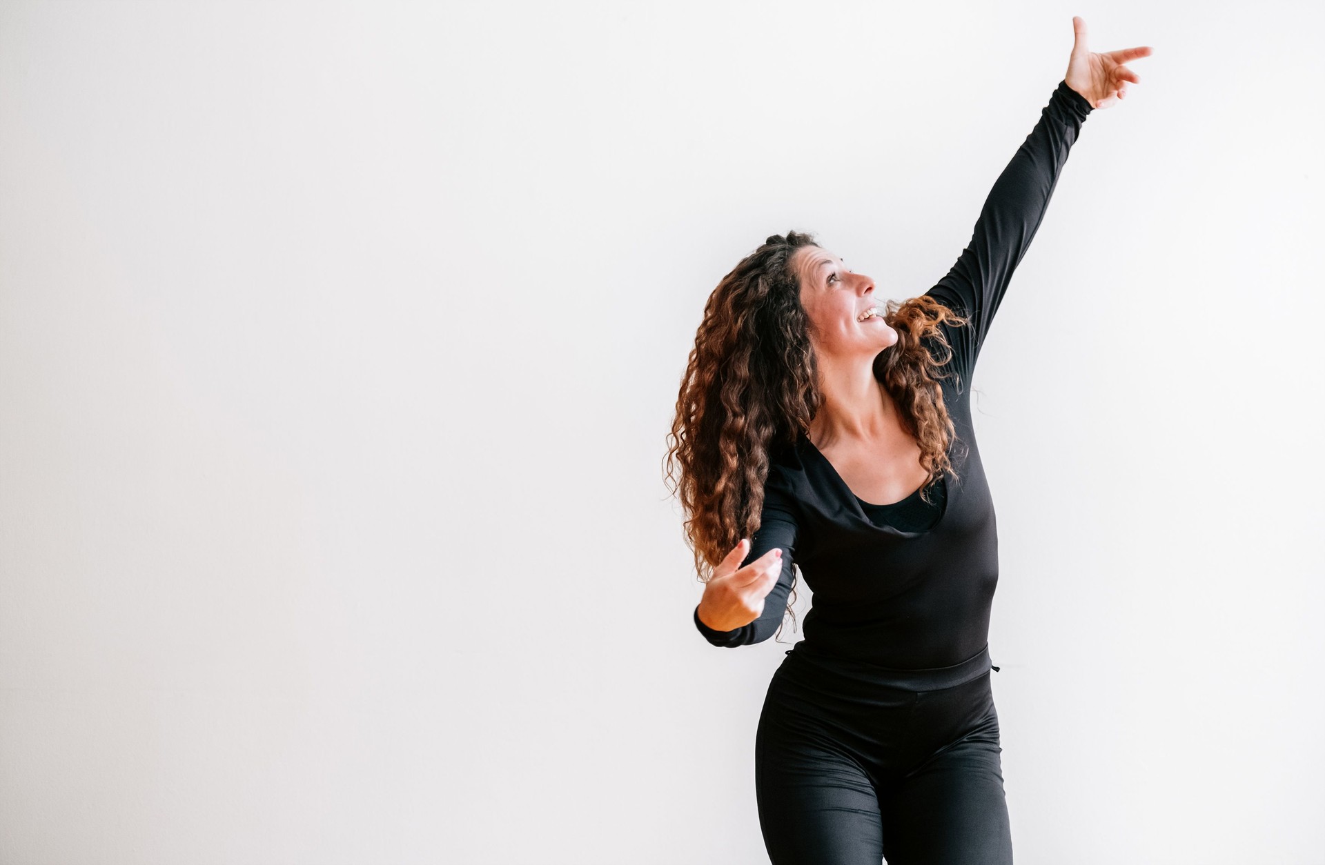Joyful woman in black outfit captured mid-dance in bright room arms emotionally raised. Her curly hair flows freely and her face radiates happiness and energy embodying essence of movement and freedom
